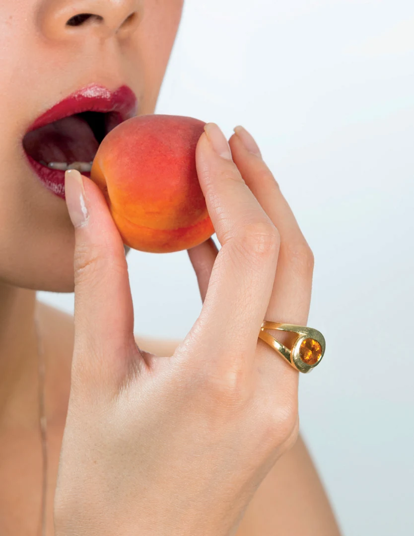 Bague Spinelle de couleur portée à la main dans un portrait du dossier de presse, avec un fruit orangé